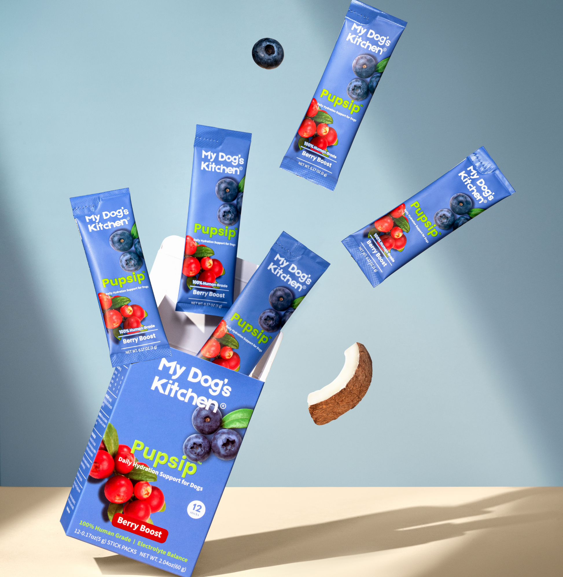 Stylized photo of PupSip Berry Boost hydration mix: a box of Berry Boost surrounded by several individual PupSip stick packs floating above, with a coconut slice on a light blue background, emphasizing natural, fun hydration for dogs